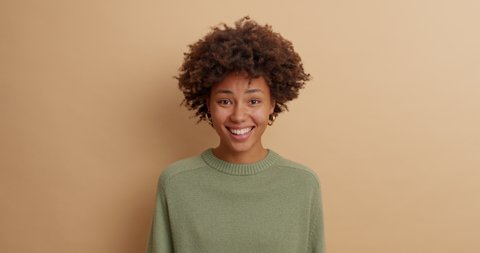 Curious dark skinned woman asks what and nods head with agreement says thats true agrees with you smiles broadly shows white perfect teeth dressed in casual jumper isolated over beige background