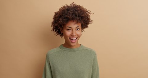 Happy flirty young woman with Afro hair winks eye and looks joyfully at camera expresses joy and satisfaction dressed in casual sweater isolated over brown background. Face expressions concept