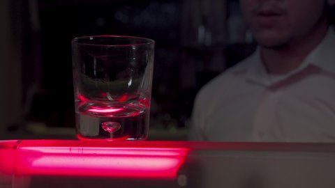 Professional Asian barman pouring liquor into rocks glass with ice ball on counter bar for serve in nightclub. Male mixologist bartender preparing alcohol cocktail drink with liquor shelf background