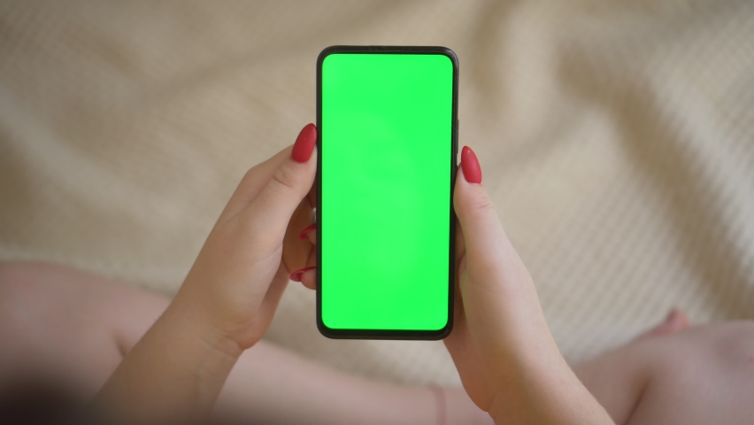 Back view over shoulder of young girl using smartphone with green screen sitting down on bed while chatting with friend.