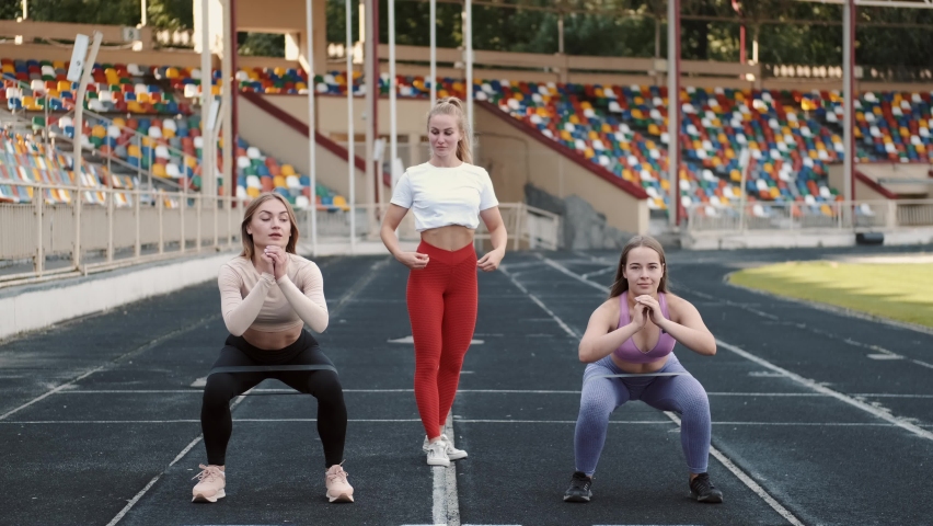 Two athletic brunette women doing exercise outdoors with the help of coach. Attractive athlete girls in stylish sportswear squatting with rubber band on stadium.