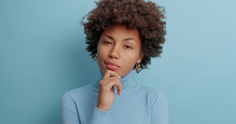 Serious dark skinned woman with thoughtful expression keeps finger on face concentrated somewhere pensively has curly bushy hair dressed in poloneck isolated over blue background. Thoughts concept