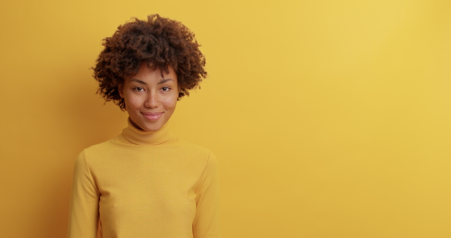 Pretty curly haired woman smiles broadly and turns head away looks on blank space over yellow wall reads pleasant announcement sees awesome advertisement rejoices sale dressed in casual wear Royalty-Free Stock Footage #1061620651