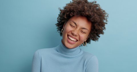 Happy emotions and feelings concept. Positive good looking Afro American woman smiles broadly has upbeat mood shows white perfect teeth dressed in casual jumper isolated over blue background