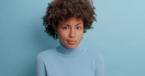 Uncertain clueless woman with Afro hair hesitates about something says no and feels puzzled dressed in casual turtleneck isolated over blue background. Indecisive questioned young female poses indoor