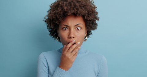Astonished embarrassed dark skinned young woman stares in disbelief covers mouth and has frightened expression stands speechless dressed casually isolated over blue background. Shock and stupor