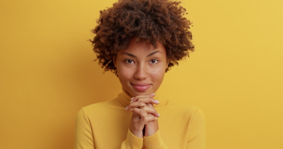 Glad cheerful woman with curly hair bites lips smiles broadly keeps hands pressed together near chin expresses happiness and joy awaits for something very good models against yellow background Royalty-Free Stock Footage #1061620765