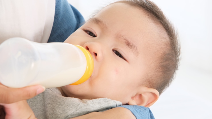 Asian mother holding little cute baby girl in arms and feeding baby from milk bottle on bed,food and drink for growth adorable infant  by care famliy.