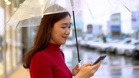 B-Roll 4K footage showing a beautiful young Asian woman standing beside the street in downtown during the rain with an umbrella, cheerful Asian woman portrait in footage. Woman using smartphone.