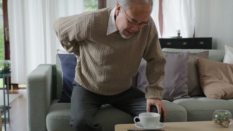 Asian elderly senior male grandpa sitting at sofa couch at home living room on quarantine in concept healthcare, chronic health issue or low back pain in retired older people.