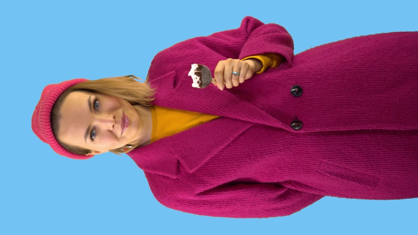 Vertical shot. Pretty Woman Bites Ice Cream Stick in Chocolate Glaze, Looks at Camera, Smiling and Showing Thumb Up Gesture. Woman Dressed in Knitted Hat, Red Coat on Blue Background in Studio. | Shutterstock HD Video #1061624743