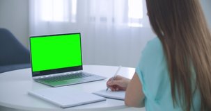 Woman sits at desk in bedroom, she looks at laptop green screen and talks to someone over internet video communications, sometimes taking notes in notebook. Close-up