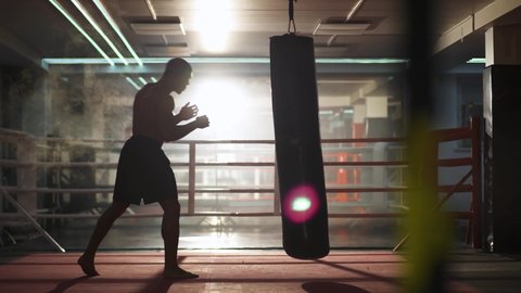 Kickboxing, athletic man fighter trains his punches, beats a punching bag, training day in the boxing gym, strikes with his feet, back lighting.