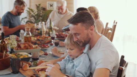 Cute little boy sitting on knees of cheerful father and playing with wooden toy plane while having fun at holiday dinner with family - Βίντεο στοκ