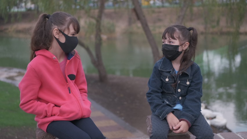 Children meet in quarantine. Nice two little girls in medical masks chatting in the city park by the water.
