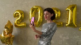 Happy new year 2021. Young pretty woman have a video call holding a phone on New Year's Eve. Female wearing evening dress at glamorous party with golden number-shaped balloons on the wall.