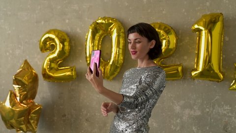Happy new year 2021. Young pretty woman have a video call holding a phone on New Year's Eve. Female wearing evening dress at glamorous party with golden number-shaped balloons on the wall. Arkivvideo