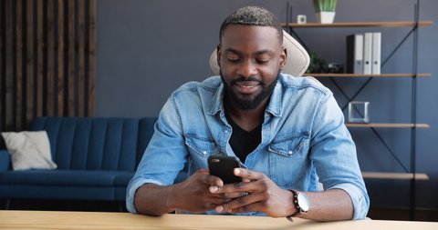 Young African American Man is sitting on the Desk in Living Room, browsing his Smartphone. Handsome Male is texting Messages, chatting in Social Media or Date Apps, having Fun. Positive Emotions.