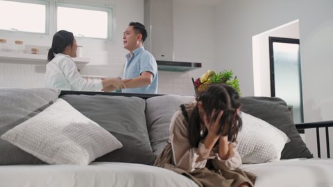 Asian girl kid sitting and crying on bed while parents having fighting or quarrel conflict at home. Child covering face and eyes with hands do not want to see the violence. Domestic problem in family.