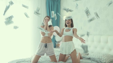 Two young beautiful smiling women in white pajamas. Sexy models in bed in hotel room.They going crazy at bachelorette party. Female throwing out falling money dollars banknotes