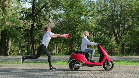 A guy in a white T-shirt with a gift in a red box catches up with a girl on a red electric scooter.