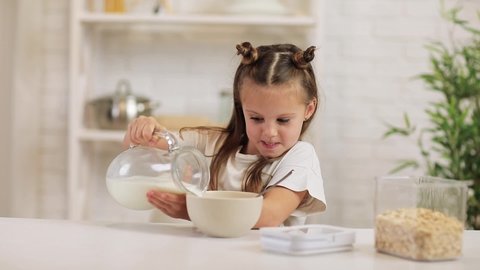 child pours milk into a bowl of cereal in the kitchen. cute little girl eating breakfast: cereal with the milk. healthy breakfast