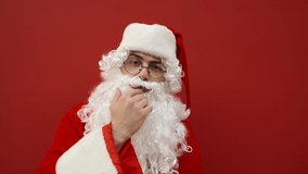 Funny Santa taps his finger on the camera and points aside at the copy space. Santa at Christmas asks to look at the recommendation on the background of the red wall.
