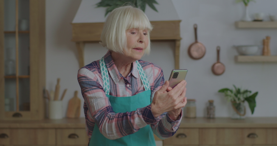 Cheerful senior blonde woman singing looking at mobile phone in hands at wooden kitchen. Elderly female having fun staying at home. Grandmother using cell phone. | Shutterstock HD Video #1061648080