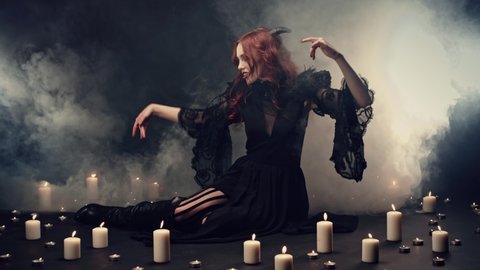 Red-haired witch sitting on floor in smoke, making energetic hand gestures to call demons. Magical ritual to invoke Satan. Cosplay, halloween costume. Concept of supernatural, evil, darkness, gothic.
