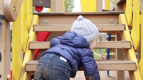 Funny little boy in a warm jacket and hat crawls, climbs and climbs up a wooden ladder on the Playground in the Park. The kid got all dirty playing in the street. The concept of a happy childhood