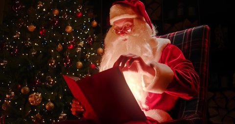 santa claus sits near a decorated christmas tree and looks into a large glowing book adjusting his glasses: stockvideo