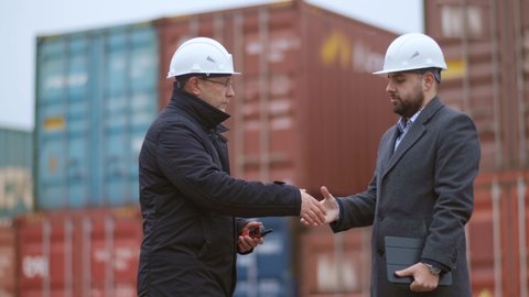 Two engineers in white helmets supervise loading at a container yard for delivery to a cargo ship for global import and export, supporting the logistics concept. Shake hands.