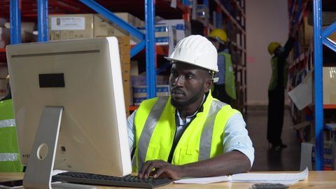 african warehouse manager sitting at desk using computer check inventory items online at distribution warehouse. black man worker  in Hard Hat typing keyboard entering data document  in workplace