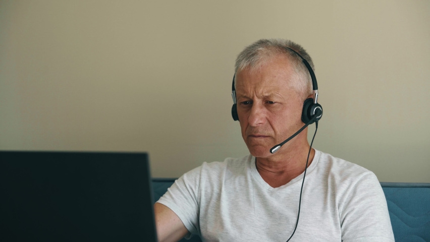 An elderly man with gray hair wearing headphones sits in front of a computer and participates in online negotiations. The concept of remote work during a pandemic and isolation.