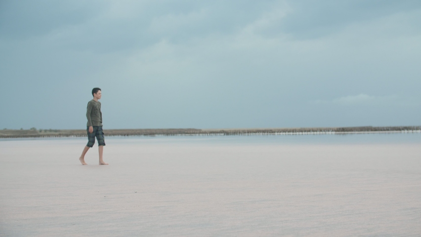 Gorgeous view of an active brunet guy in a dark T-shirt and long shorts strolling on a sandy sea beach and looking around on a drizzling day in autumn.