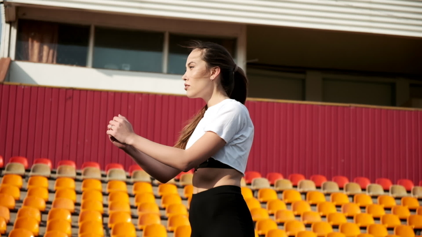 young fit brunette female in sportswear does sports exercises at stadium. slim asian attractive woman in activewear doing squats outdoors. healthy lifestyle, fitness training, workout on sunny day