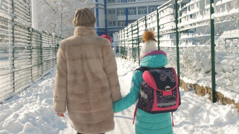 Mom in a fur coat and daughter in the jacket together go to school on a cold winter morning.