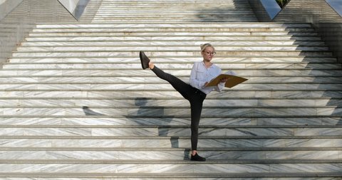 A beautiful girl, standing on the street stairs, revises documents in a yellow folder, lifts her right leg up. Flexibility in a work with papers and documents.