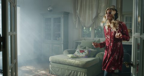 Cinematic shot of young carefree happy attractive blond woman wearing red dress and headphones is having fun to listening to the music, dancing crazy and jumping on sofa in living room at home.