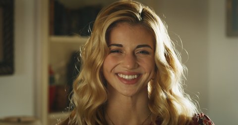Cinematic shot of young attractive happy woman with long blond hair  and piercing in her eyebrow is smiling in camera in a living room at home. Concept: non stereotyped people, identity and diversity.