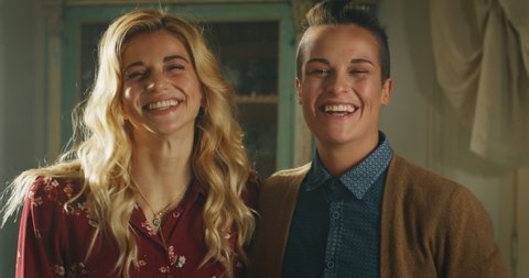 Cinematic shot of young happy homosexual female gay and transgender man couple is enjoying time together, kissing and smiling in camera at home. Concept of gender expression, identity and diversity.