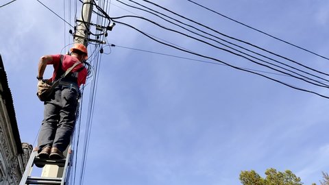 Skilled electrician in helmet fixes wires standing on ladder near high pole against blue sky on summer day backside view