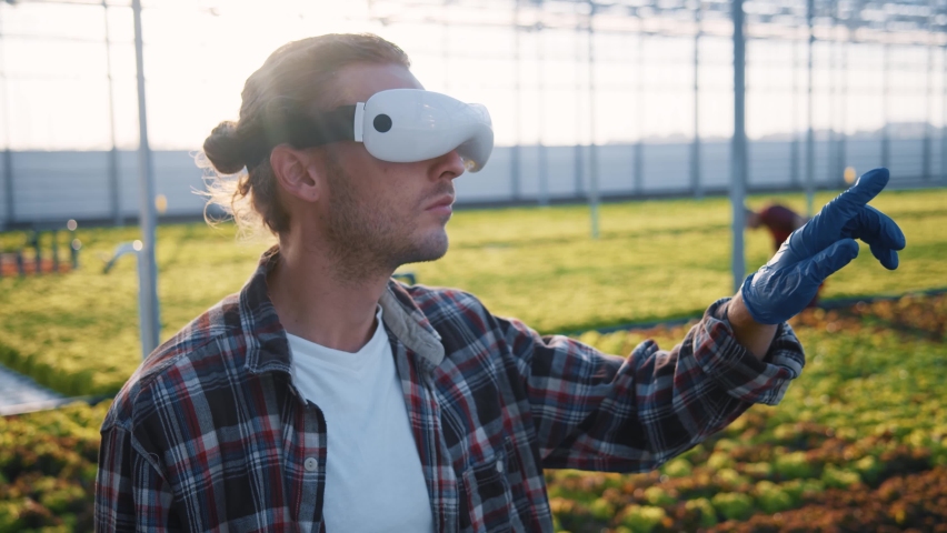 Bearded young caucasian worker wearing VR headset entertaining virtual agronomy scientific hologram managing quality system programming irrigation farming at greenhouse. Royalty-Free Stock Footage #1061674108