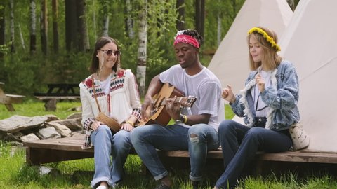 Trio of multi-ethnical friends sitting on bench at summer campsite listening and dancing to live guitar playing of young African American