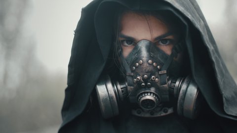 Close-up portrait human of future. Woman in grunge clothes hooded mantle and gas mask standing in forest look into camera. Dystopian concept, female survivor. Post apocalyptic world. Cyberpunk soldier