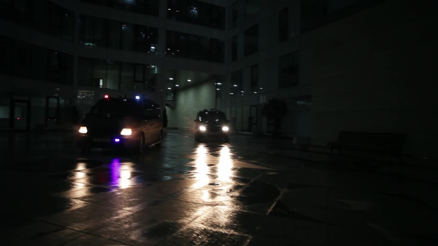 Two police vans with flashing lights quickly arrive at the crime scene at night. It is raining heavily, reflecting the glare on the wet asphalt.