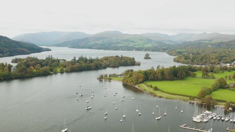 aerial shot from a drone of lake windermere in the foreground there are fields and the small town of bowness in the background you can see the mountains and hills of the Lake District