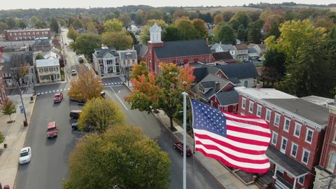 Manheim , PA / United States - 10 18 2020: Close-up aerial of US American flag waving above Main Street in Small Town America square. Establishing shot as traffic passes. Autumn fall foliage at church