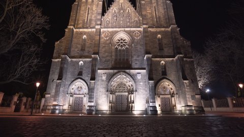 The illuminated entrance and front side of the gothic stone medieval church of saint Peter and Paul with its two spires at Vyšehrad,Prague,Czechia,at night,on an empty paved square with lanterns,tilt.