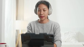 Asia businesswoman wear headphone using tablet talk to colleagues about plan in video call while smart working from home at bedroom. Self-isolation, social distancing, quarantine for corona virus.
